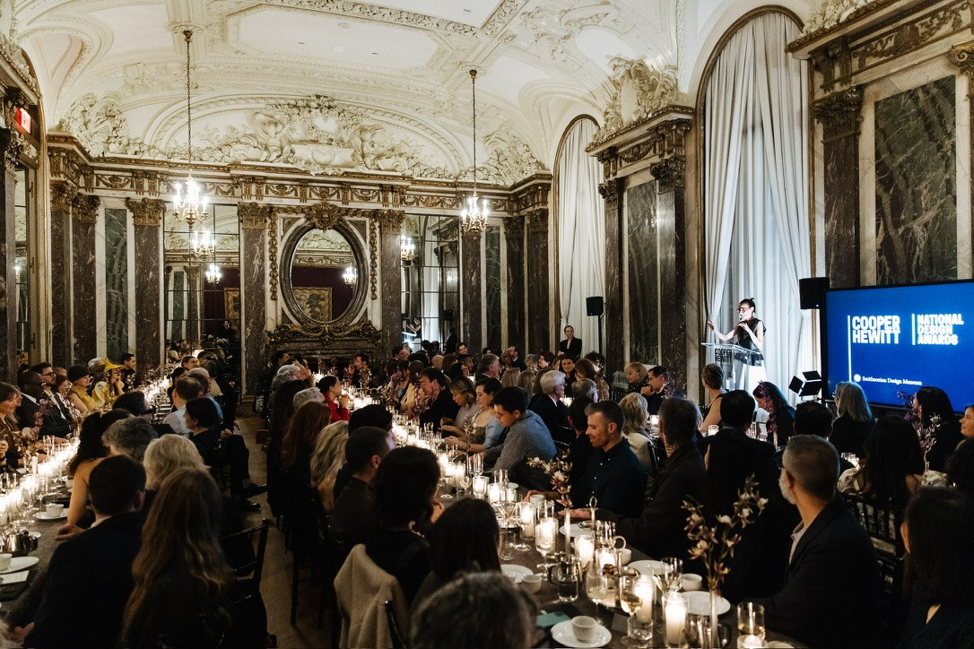 The National Design Awards dinner in an ornate hall with marble columns and chandeliers, long candlelit tables of guests facing a speaker at a podium beside a Cooper Hewitt screen.