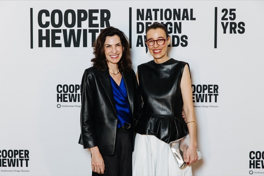 Jules Sherman, in a black leather jacket and blue blouse, smiling beside another guest in front of the Cooper Hewitt National Design Awards backdrop.