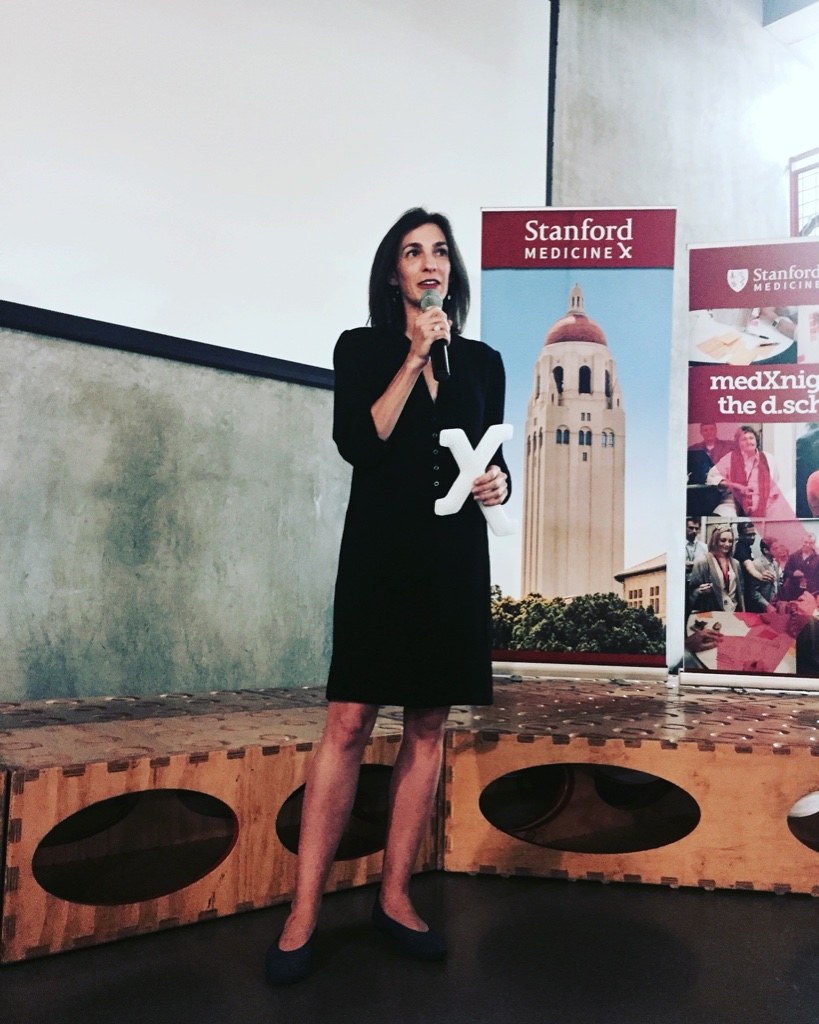 Jules Sherman speaking into a microphone and holding a white X-shaped award, in front of Stanford Medicine X banners.