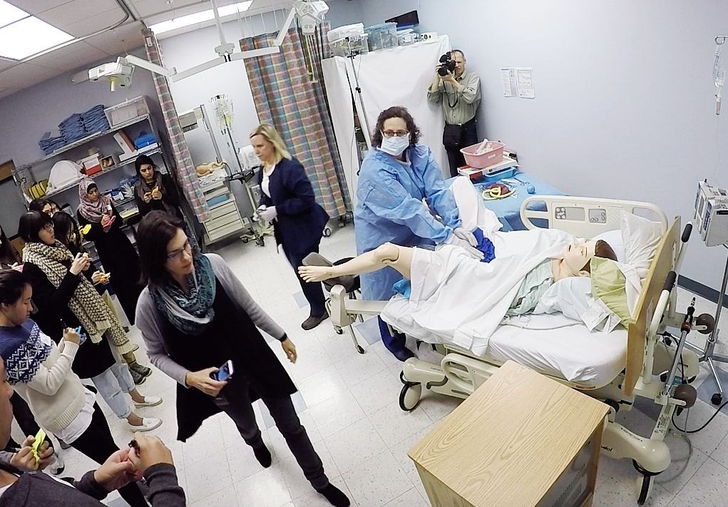 Jules Sherman guides students taking notes as a clinician in a gown works with a birthing mannequin on a hospital bed during a simulated delivery.