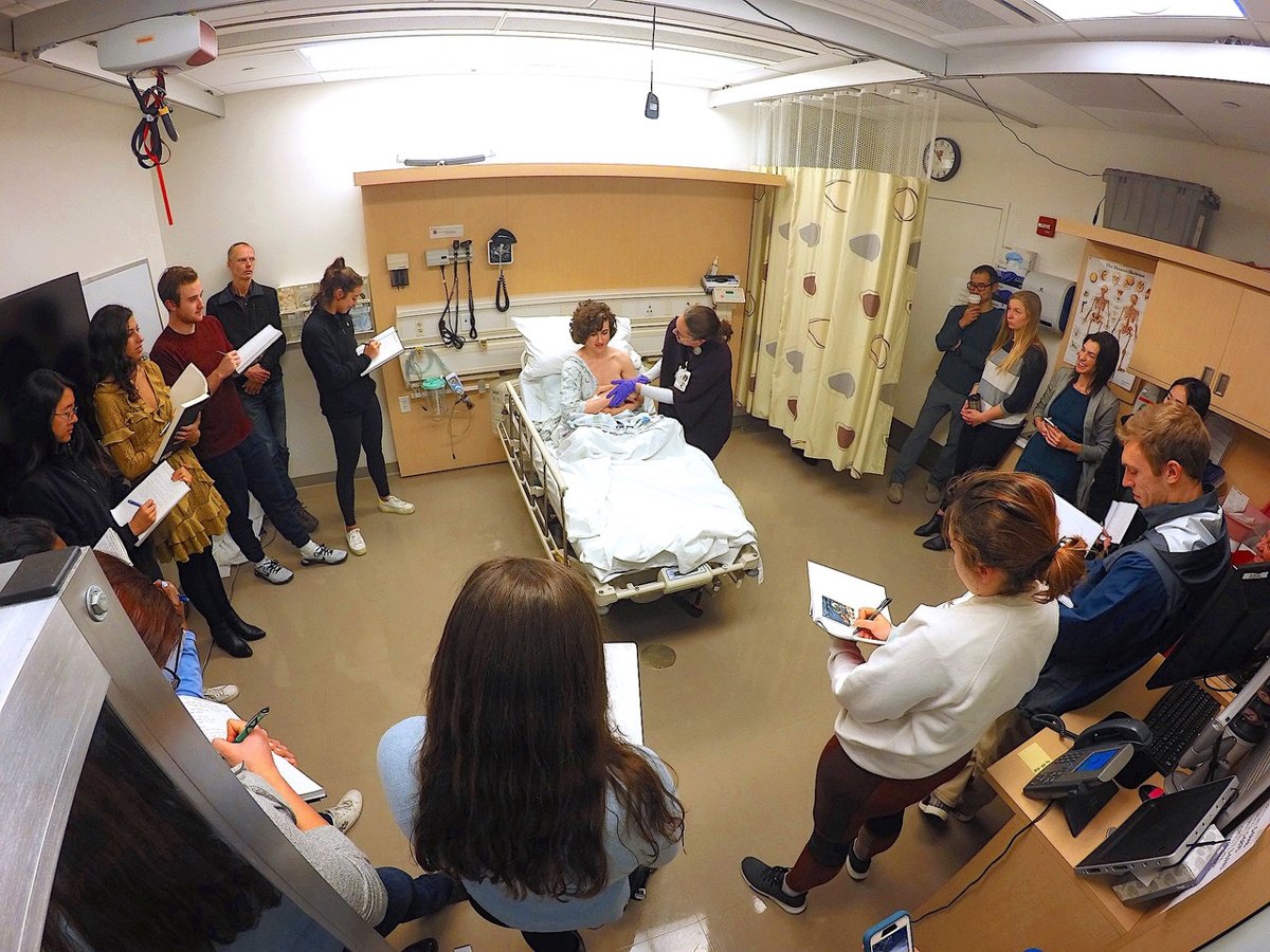 Students with notebooks stand around a hospital bed in a simulation room, observing an actor playing a new mother with a clinician beside her.
