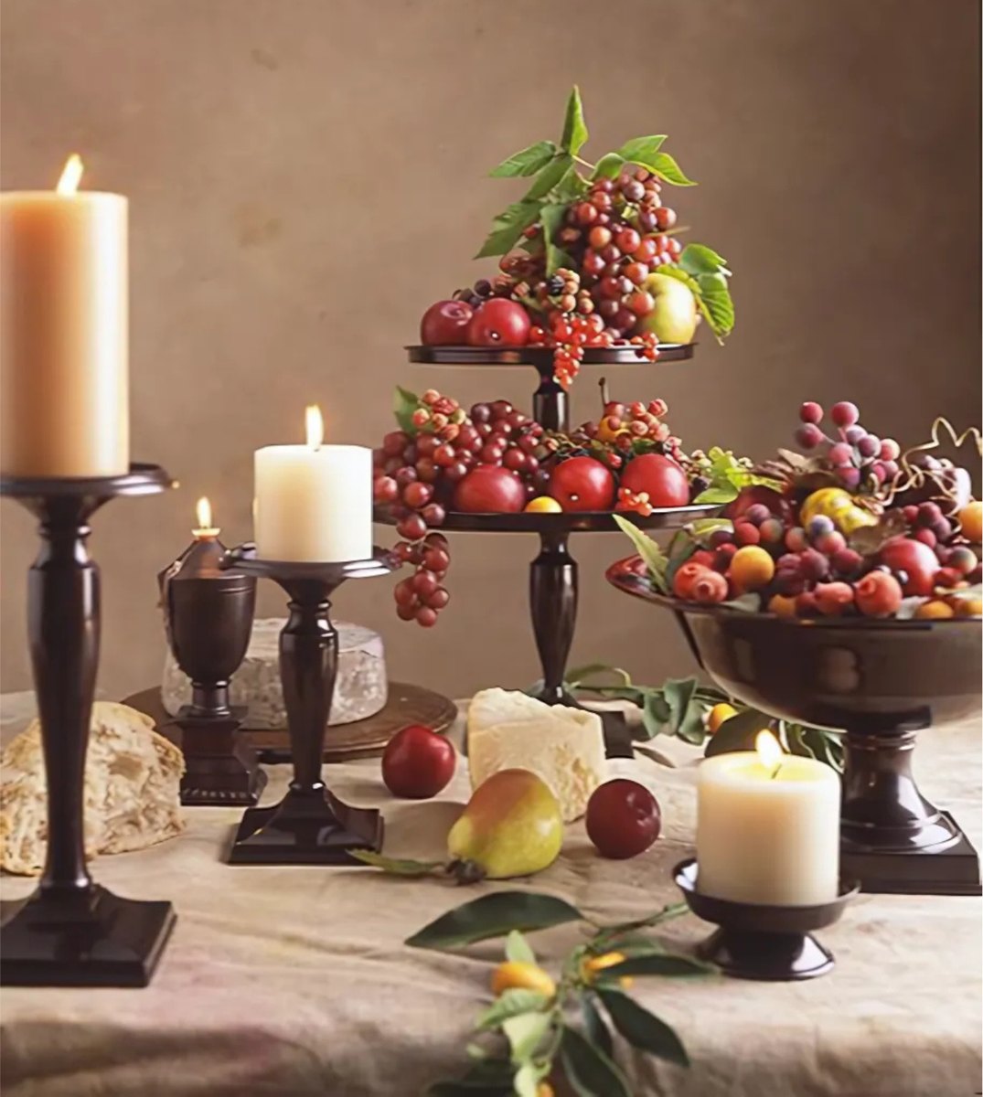 Cream pillar candles on dark candlesticks and stands, set on a linen-covered table among tiered dishes of grapes, berries, pears and cheese.