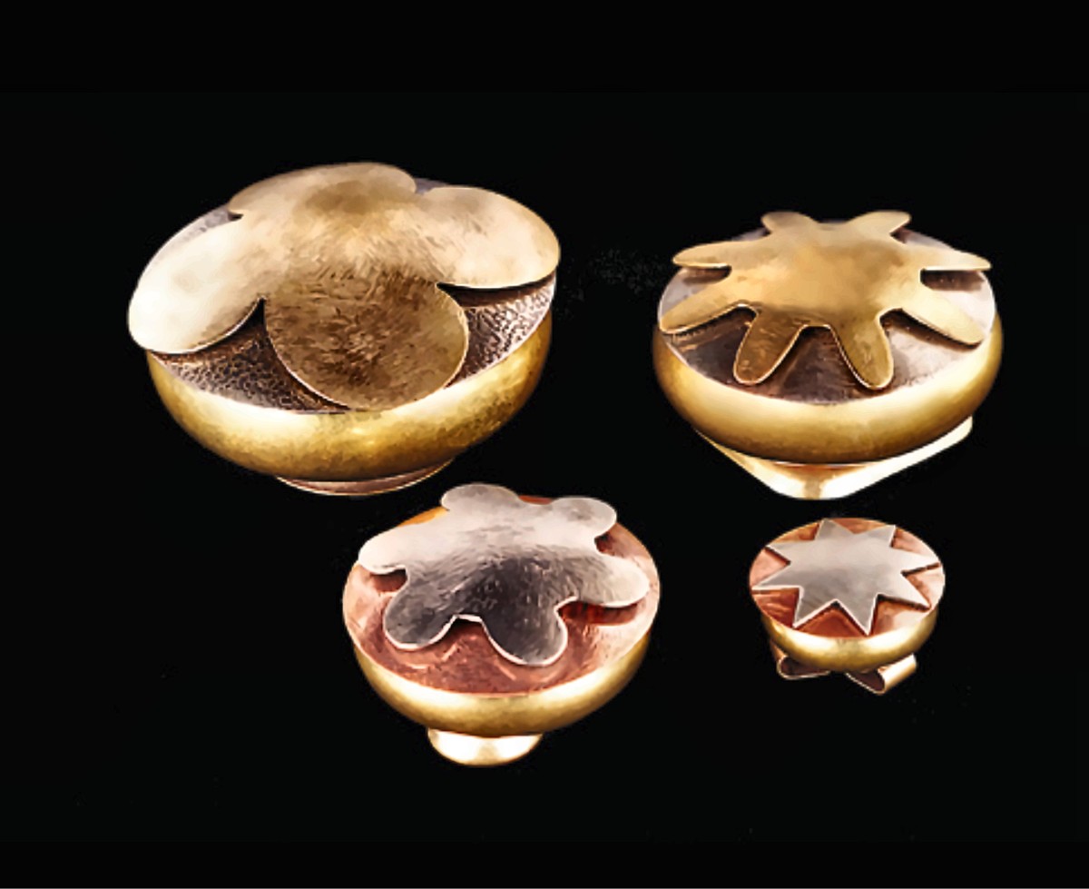 Four round lidded boxes in brass, silver and copper, of different sizes, each lid topped with a cut-metal flower, star or starburst shape, on black.