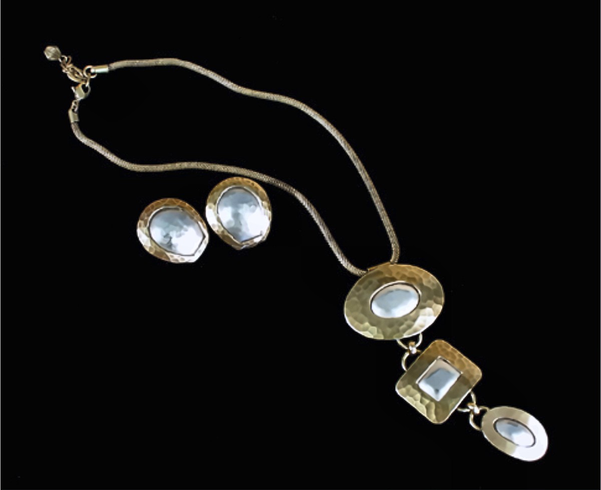 A hammered brass necklace on a snake chain, its pendant three linked plaques, an oval, a square and a small oval, each set with a polished silver center, beside matching oval earrings, on black.
