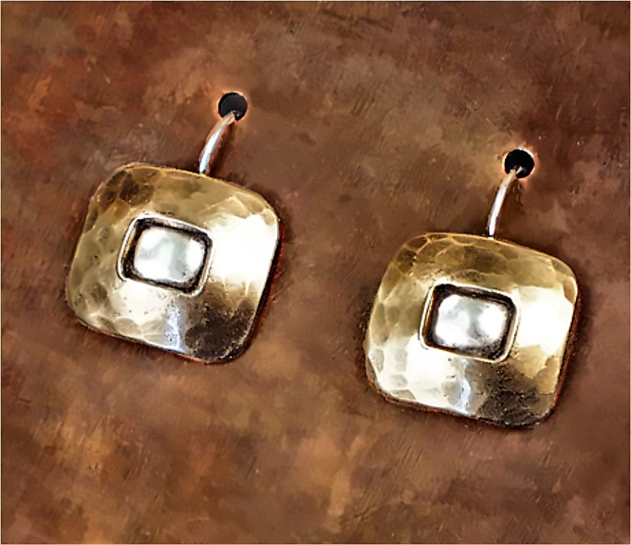 A pair of square hammered brass drop earrings, each with a raised polished silver center, on hooks against a mottled copper ground.