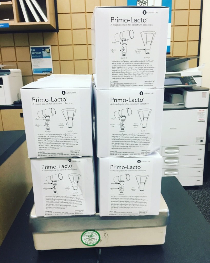 A stack of five Maternal Life Primo-Lacto boxes, printed with drawings of the adapter, collection cup and hand-expression funnel, on a shipping scale at a post office.