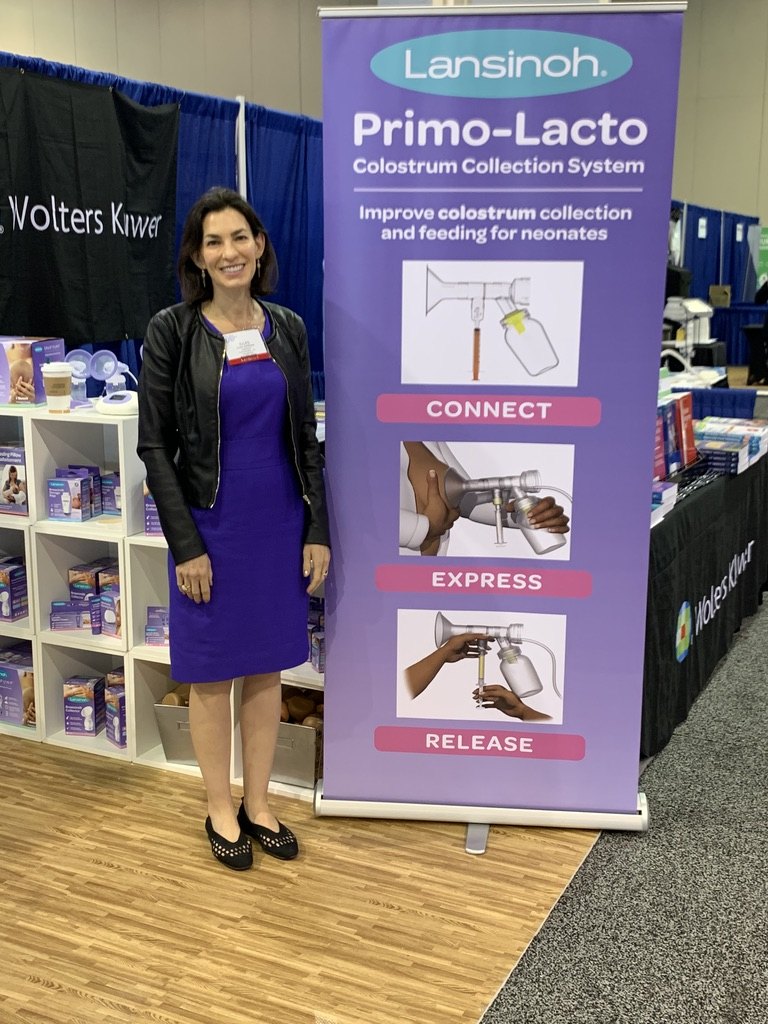 Jules Sherman standing beside a Lansinoh Primo-Lacto Colostrum Collection System banner, showing the connect, express and release steps, at a trade show booth.