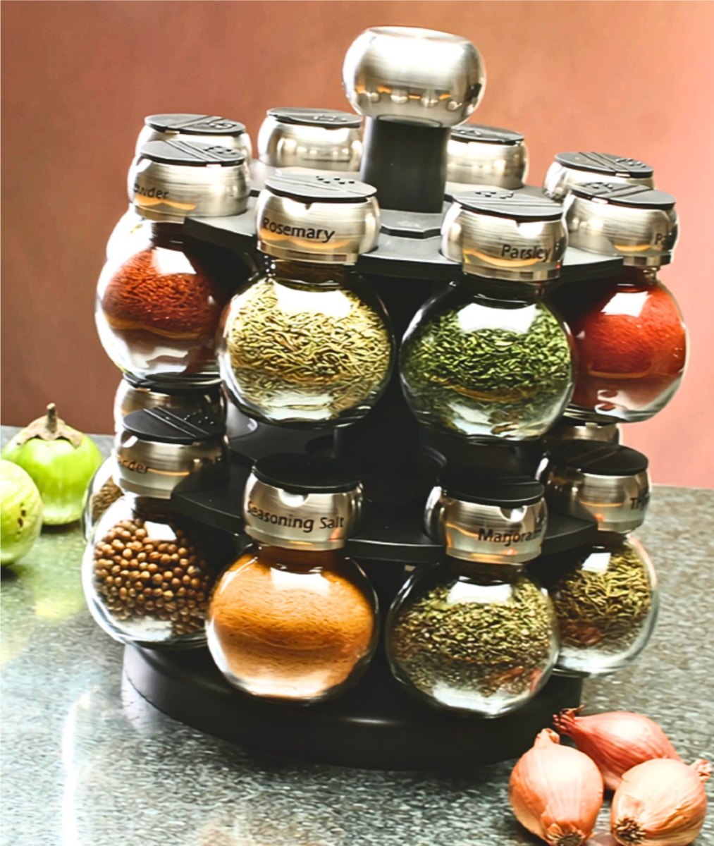 A black two-tier rotating spice rack holding round glass jars of colorful spices with stainless steel lids, on a granite counter with shallots and tomatillos.