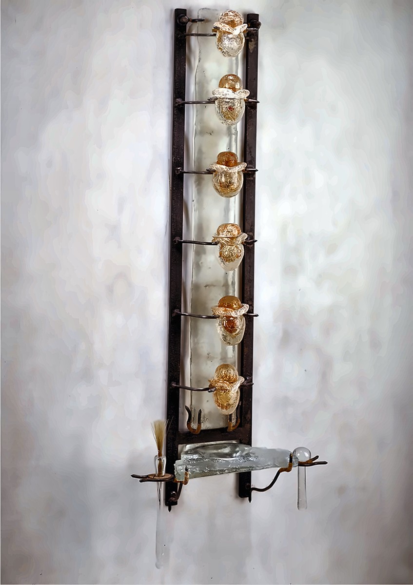 A tall, narrow wall piece with a dark metal frame holding a column of amber glass forms above a small glass shelf with a brush and a glass rod.