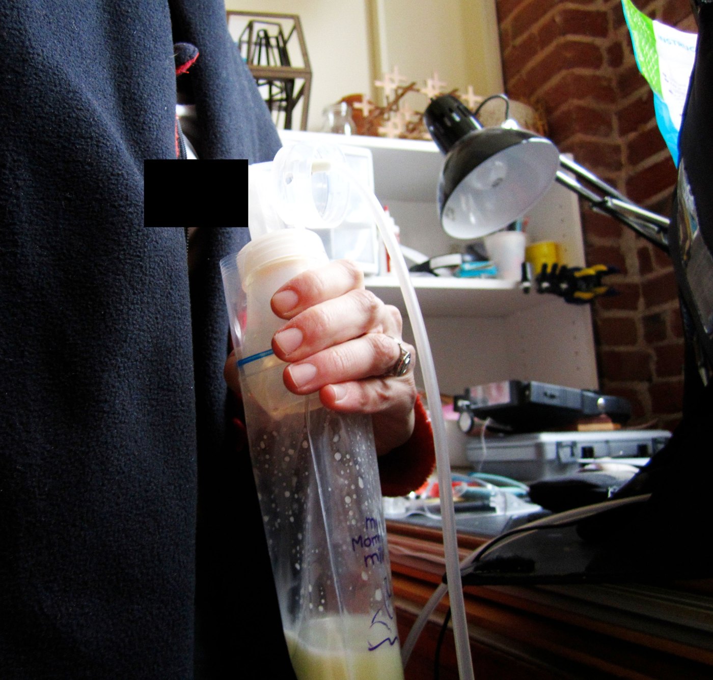 A hand holding the breast pump collection piece attached to a milk storage bag, with tubing running to the pump, in a studio.