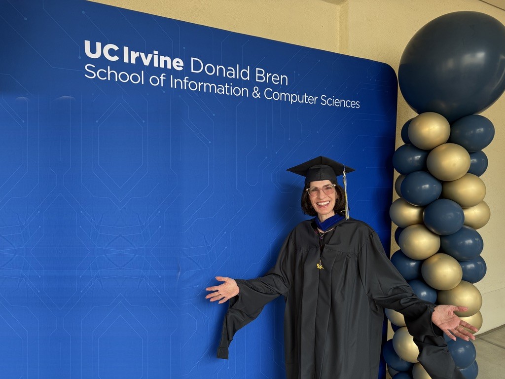 Jules Sherman in cap and gown, arms open and smiling, in front of the UC Irvine Donald Bren School of Information and Computer Sciences backdrop beside a column of blue and gold balloons.