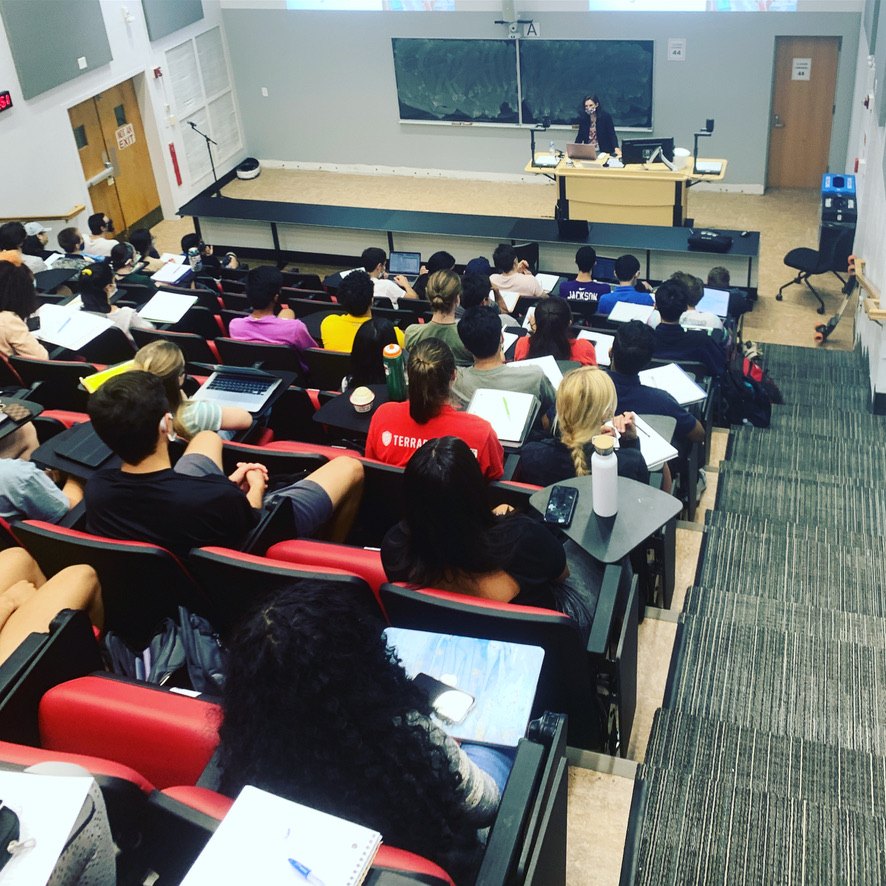 Jules Sherman lecturing at the front of a raked lecture hall at the University of Maryland, seen from the back rows of students with laptops and notebooks.