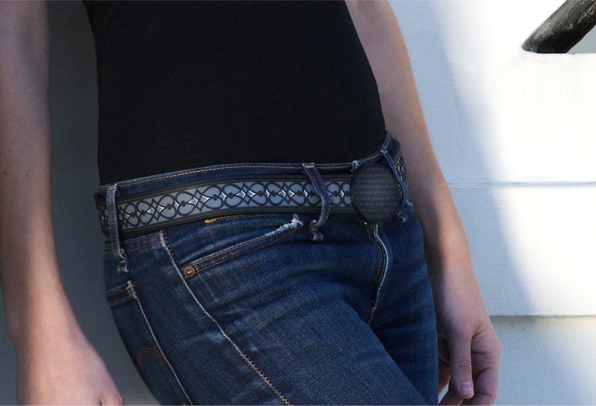 A black belt with a gray interlocking pattern and a round patterned buckle, worn through the loops of dark blue jeans.