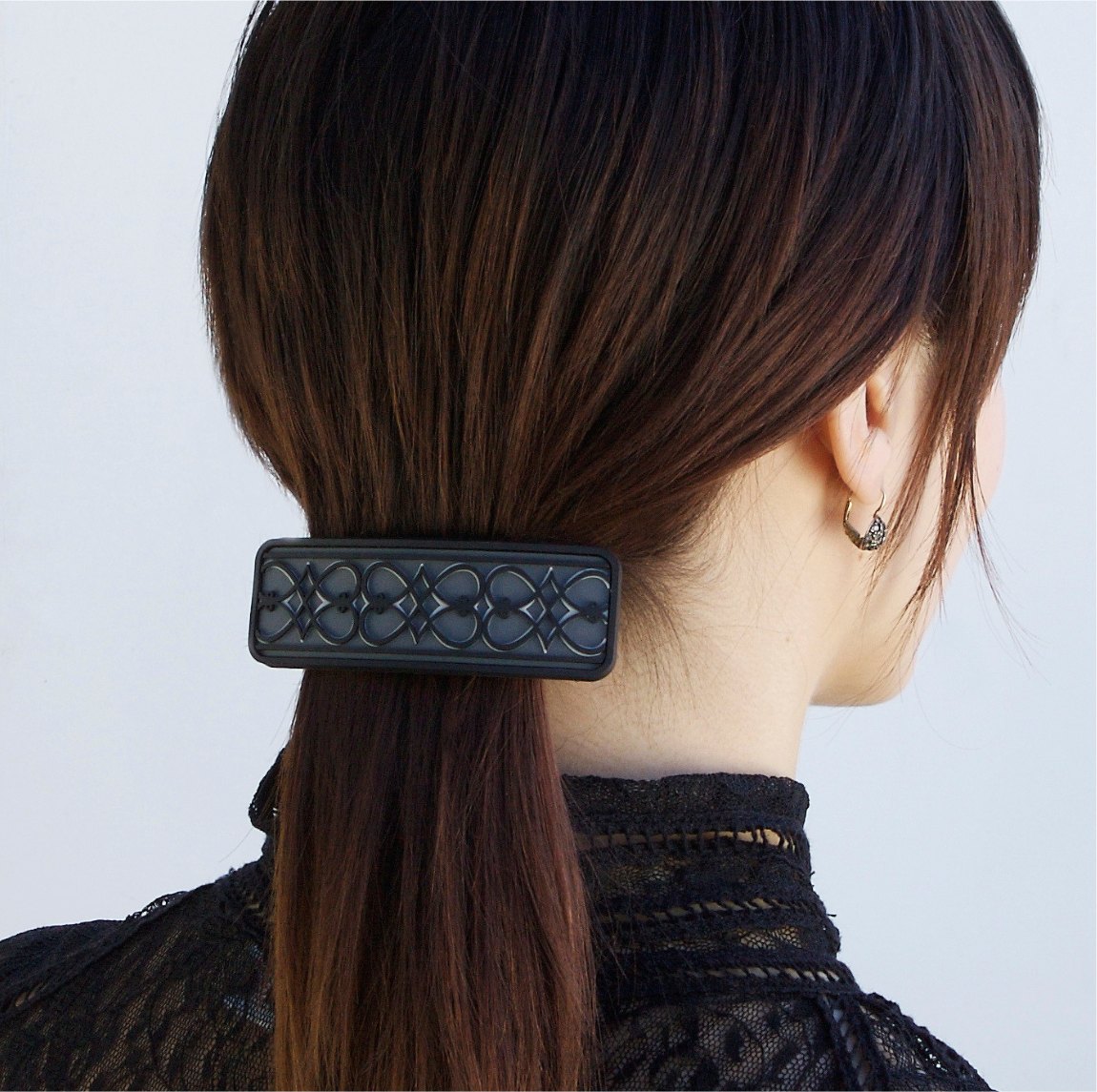 A woman seen from behind with her hair in a low ponytail, held by a black rectangular barrette molded with an interlocking pattern.