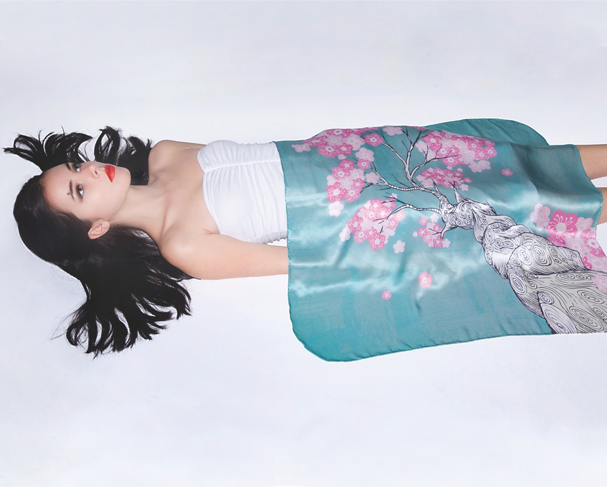 A model lying on her back on a white ground, a teal silk scarf printed with a pink cherry blossom tree draped across her body.
