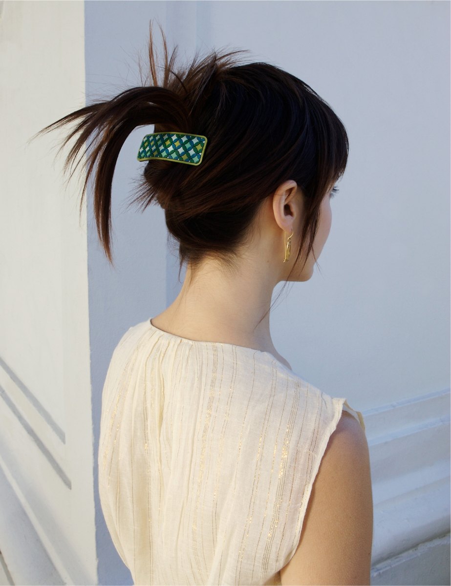 A woman seen from behind with her hair in a loose twist, held by a rectangular green barrette with a woven gold pattern.