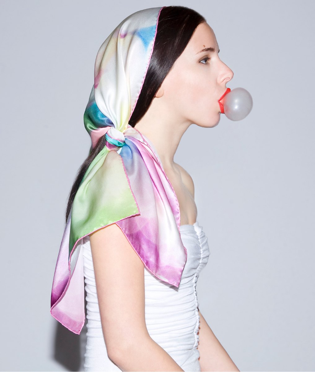 A model in profile blowing a bubble of gum, her hair wrapped in a silk scarf printed in soft pink, blue and green watercolor washes.
