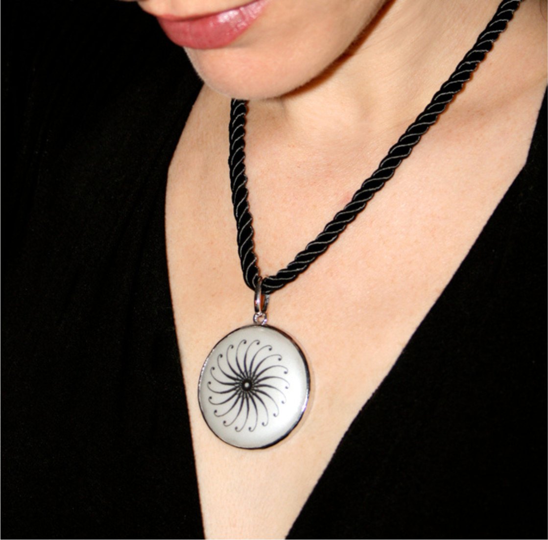 A round white pendant with a fine black spiral pattern, framed in silver and hung on a twisted black cord, worn with a black V-neck top.